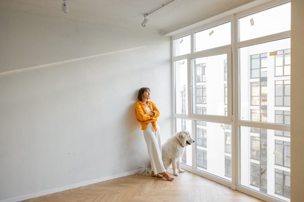 Young woman stands with her dog near the window in bright room of her new apartment. Friendship with pet and house renovation concept